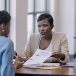 A Black woman at the desk is showing an application document to an African American woman sitting opposite her.