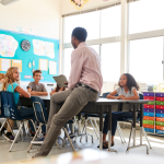 A teacher leans against a table while addressing a small group of students.