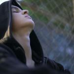 Teen wearing a hoodie leaning back on a chain link fence.