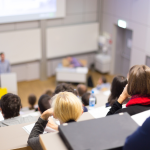 Students listening to their professor speak in a large lecture hall.