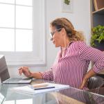 A woman sitting at a desk, grimacing and holding her hand on her back.