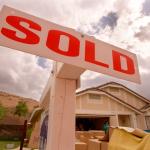 Close-up of a sign reading "Sold" in bright orange in front of a house and lawn full of boxes.