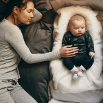 A mother sleeping next to a baby in a bassinet.