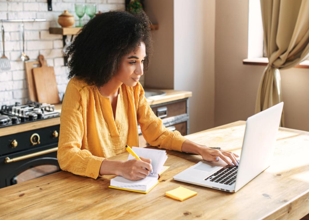 A woman wearing a yellow blouse takes notes while watching a laptop.