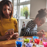 A woman checking her phone while a little girl does crafts next to her.