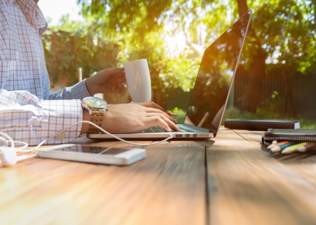 A person in a dress shirt sits outside with a laptop and a cup of coffee.