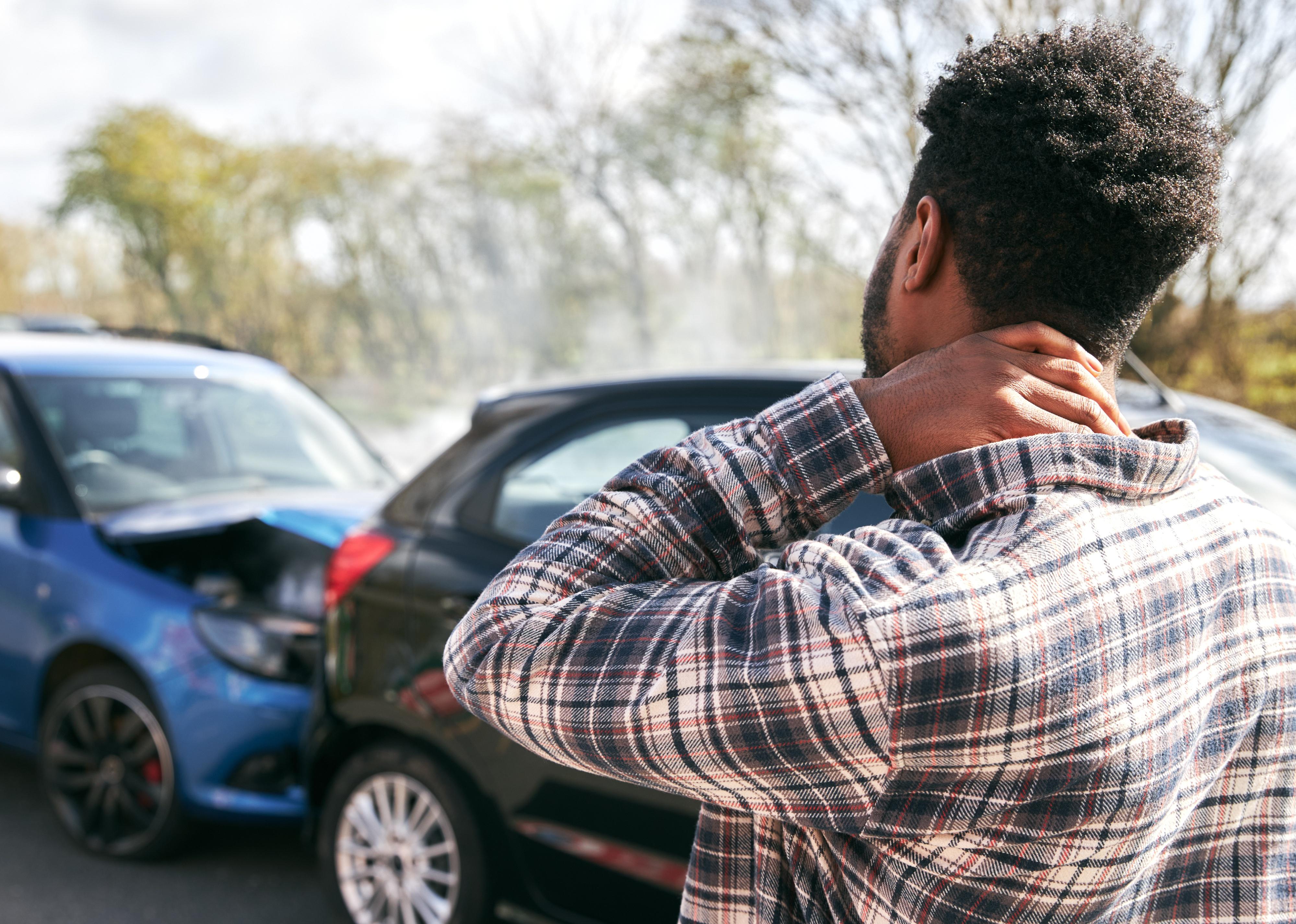 Young man rubbing neck in pain standing by damaged car after traffic accident.