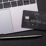 Black premium bank card lays on a silver macbook laptop computer next to the trackpad, with a shiny fancy pen on the table.