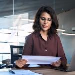 Focused young businesswoman reading financial report document and using a laptop in the workplace.
