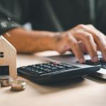 A person using a laptop and calculator with a wooden figure of a house and coins beside him.