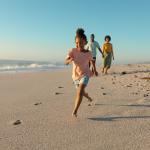 A young girl running on sand against parents walking on a beach.