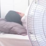 A white electric fan in the foreground in front of a person sleeping in bed, facing away.