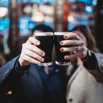 Two people holding up a pint each of stout beers to cheers, which obscures their faces from the camera in a bar.