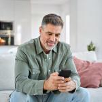 Middle aged old man using smartphone sitting on a couch with kitchen in background.