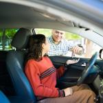 A teenage daughter inside a car receiving its keys from her proud dad.
