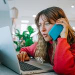 A woman talking on her smartphone while also booking flights using her laptop.