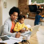 A black mother takes notes as she works at home while her young daughter sits on her lap.