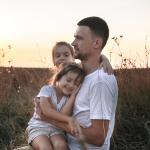 A father with her two young daughters out on a grass field during sunset.