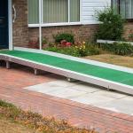 A wheelchair access ramp with green plastic surface installed at the door to a building that has a single step up to a black door.