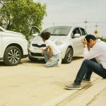 Drivers of two cars caught in a road accident inspect and sit down by a roadside in distress.