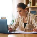 Determined-looking woman looking intently into laptop screen. She is sitting in an office with wooden bookshelves and a window behind her.