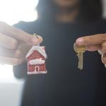 A woman holding a small figure of a Christmas home with her right hand and a house key with her left.