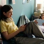 Two teenage girls relaxing on a bed and using smart devices to browse the internet.