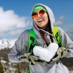 A young woman in snowboarding gear holding a heart shape made of snow.