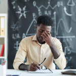 A young teacher looking concerned sitting at the desk in a classroom.