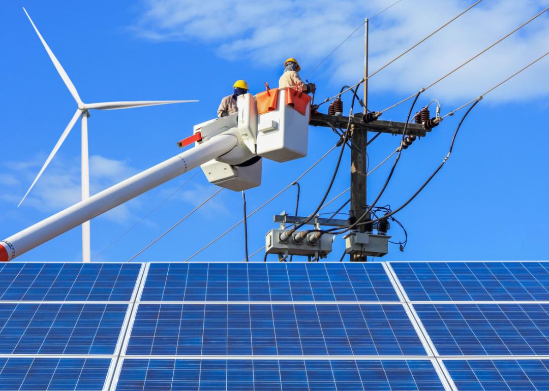 Electricians inspecting power line with solar panels and wind turbines.