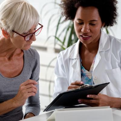 An older patient sits beside a doctor who is reviewing information on a clip board.