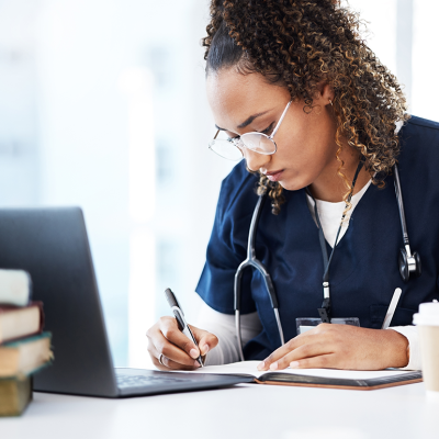 Medical student working at desk, writing in a notebook in front of a laptop, books, and a to-go coffee cup. 