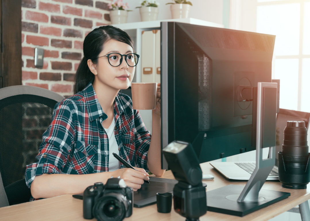 A woman with glasses looks intently at her monitor with camera equipment around her. 