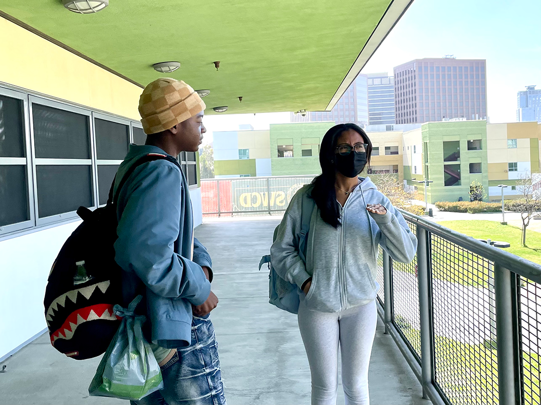 Two teeangers stand on an outdoor walkway next to a railing and under an overhanging roof.