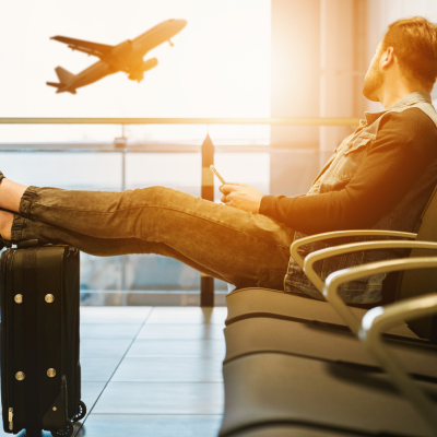 A man sits in a chair at an airport with his feet up on his carryon suitcase, watching a plane take off in the distance