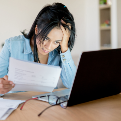 A woman seated at a desk in front of a laptop, holing paperwork and looking frustrated.