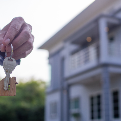 Hand holding a new house key with a blue and white house in the background. 