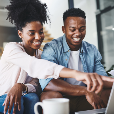 A young Black couple sit on the couch, smiling, and look at a laptop computer