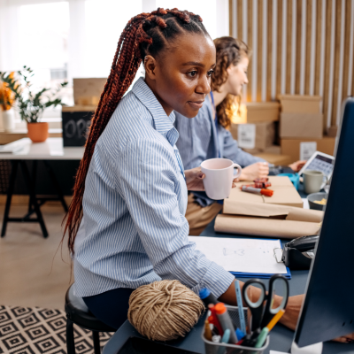 Two women sitting in a creative-looking office with plants and boxes, working on the computer and wrapping something in brown paper.