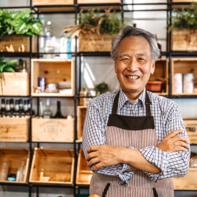 An older man smiling with his arms folded in a shop with shelves behind him.