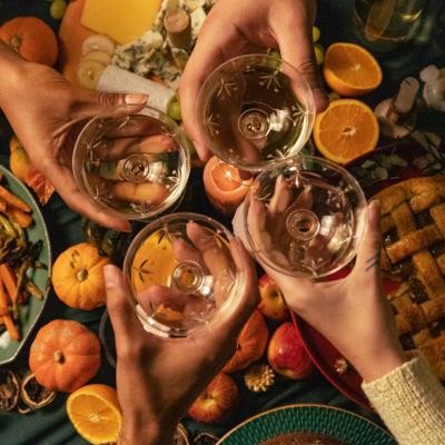 Four people toast with wine glasses over a Thanksgiving meal. Only their hands and the glasses are shown above the table.