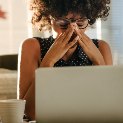 A Black woman sits in front of a laptop in a stressed posture, her hands covering part of her face