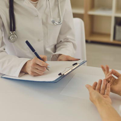 A doctor writes on a clipboard as they speak with a patient at a desk.
