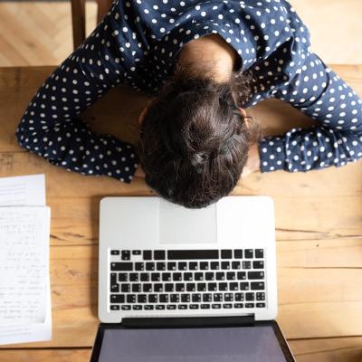 overhead view of woman resting head on desk in front of laptop computer and notes