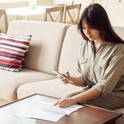 Woman holds smartphone in one hand while checking paperwork on table with the other.