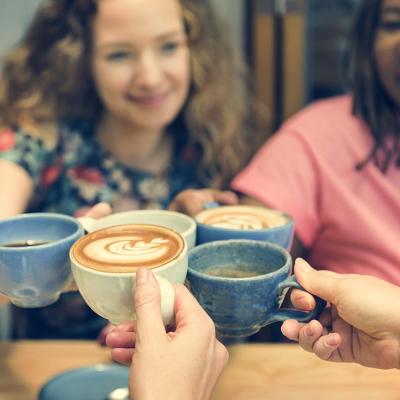 Five hands holding coffee touch their drinks together in the middle of the table with two women's face visible in background.