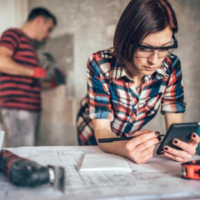 Two people in charge of overseeing a home improvement where one woman is leaning on a table with blueprints and noting with an instrument and the other is a man hammering nails on a wall.