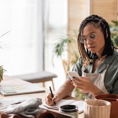 A woman with a gardening business is looking into a phone while writing down information on a notebook.