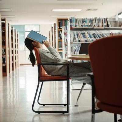 A female college student in a library with an open book over her face seemingly tired of reading.