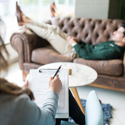 A psychotherapist writing down in a notebook while in session with a male patient who is lying on couch.